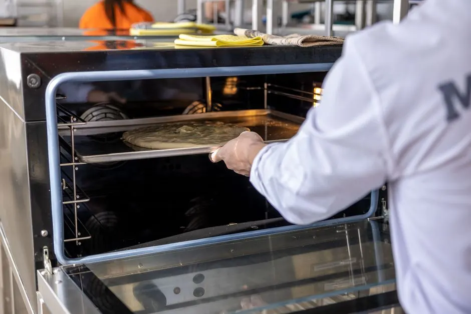 Chef places uncooked pizza into an industrial oven in a professional kitchen setting.